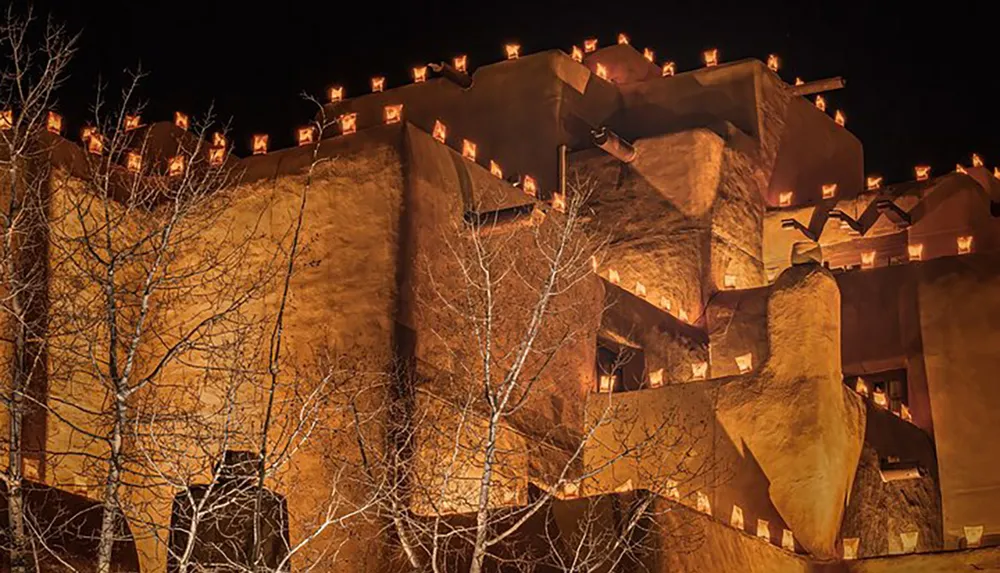 This image features the warm luminous facade of an adobe-style building highlighted by numerous small lights along its parapets set against a night sky