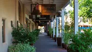 The image shows a shaded outdoor gallery lined with shops and potted plants, featuring a sign for 