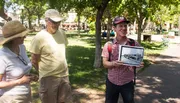 A person is holding a framed black and white photograph and showing it to two other individuals in a sunny park setting.