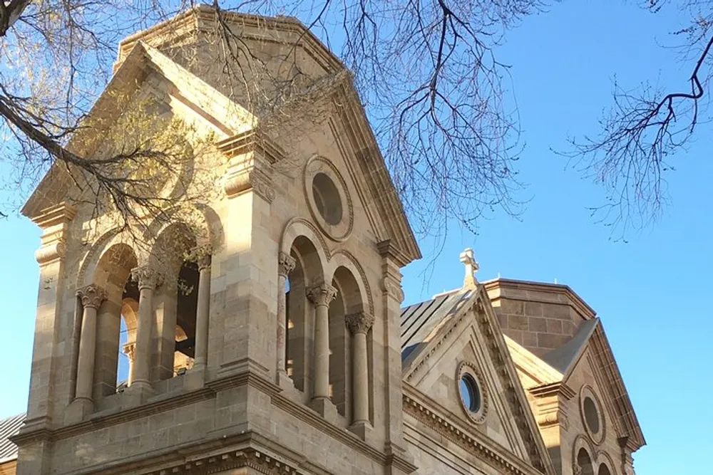 The image shows the elegant and detailed architecture of a historic building with an arched bell tower set against a bright blue sky framed by the bare branches of trees