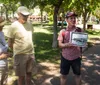 A person is showing a framed photograph to a couple in an outdoor park-like setting possibly during a tour or presentation