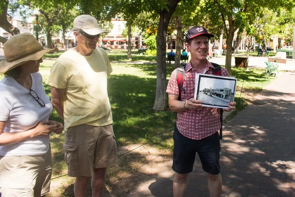 A person is showing a framed photograph to a couple in an outdoor park-like setting possibly during a tour or presentation