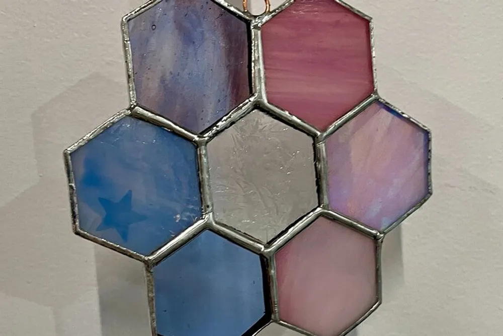 The image shows a stained glass artwork composed of hexagonal pieces in shades of blue and pink with one piece featuring a star design all joined by a metal framework