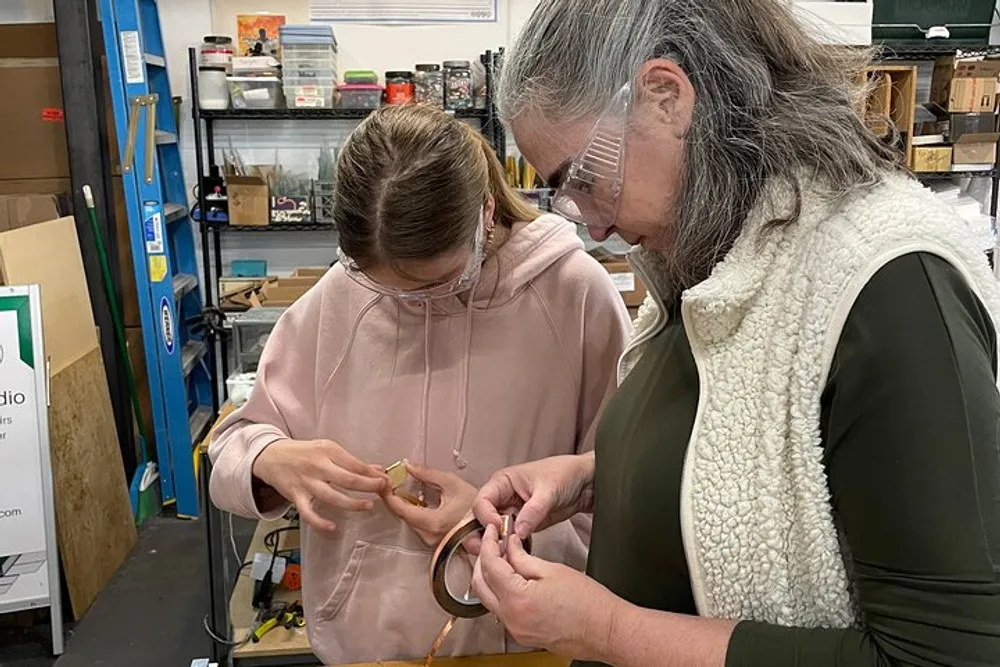 Two individuals are focused on a hands-on task one holding a ring-like object while the other appears to be guiding or teaching with both wearing safety glasses and surrounded by a workshop environment