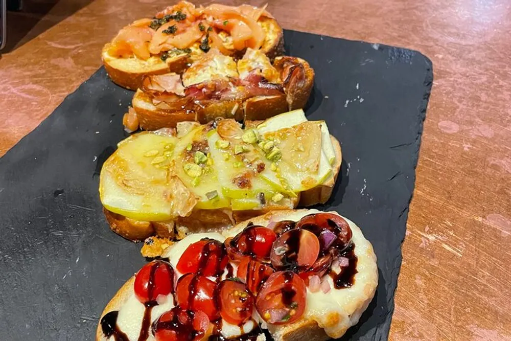 The image shows a variety of four bruschetta toppings on toasted bread artistically arranged on a slate serving board