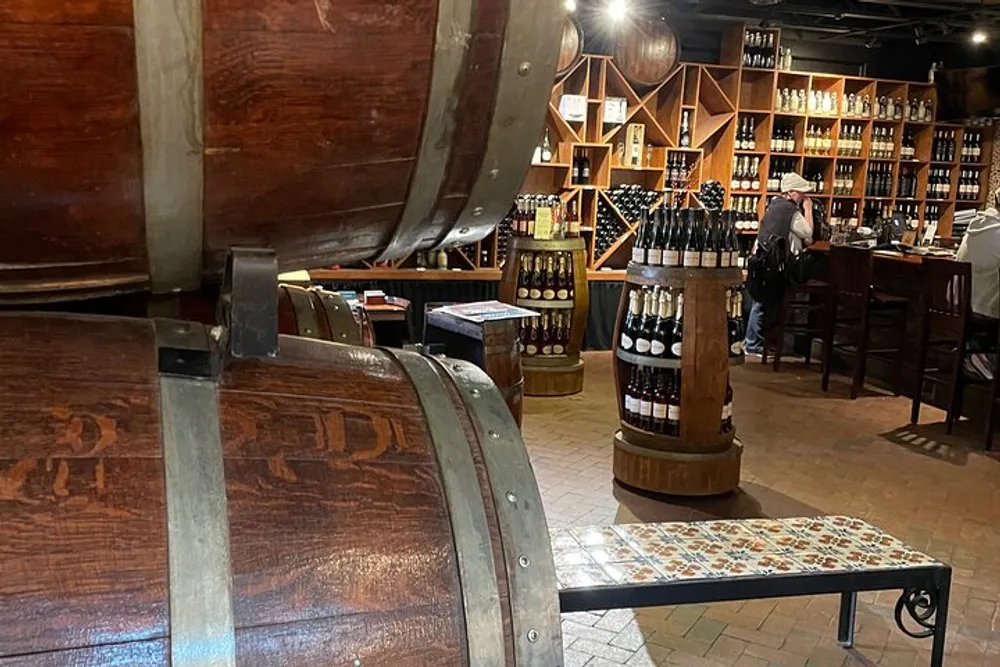 An interior view of a wine shop with wooden barrels and shelves stocked with bottles and a person in the background browsing the selection