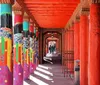 The image features a covered walkway with brightly painted artistic columns displaying vibrant patterns and colors under a terracotta-red overhang with people walking in the distance