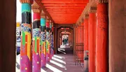 The image features a covered walkway with brightly painted artistic columns, displaying vibrant patterns and colors, under a terracotta-red overhang, with people walking in the distance.