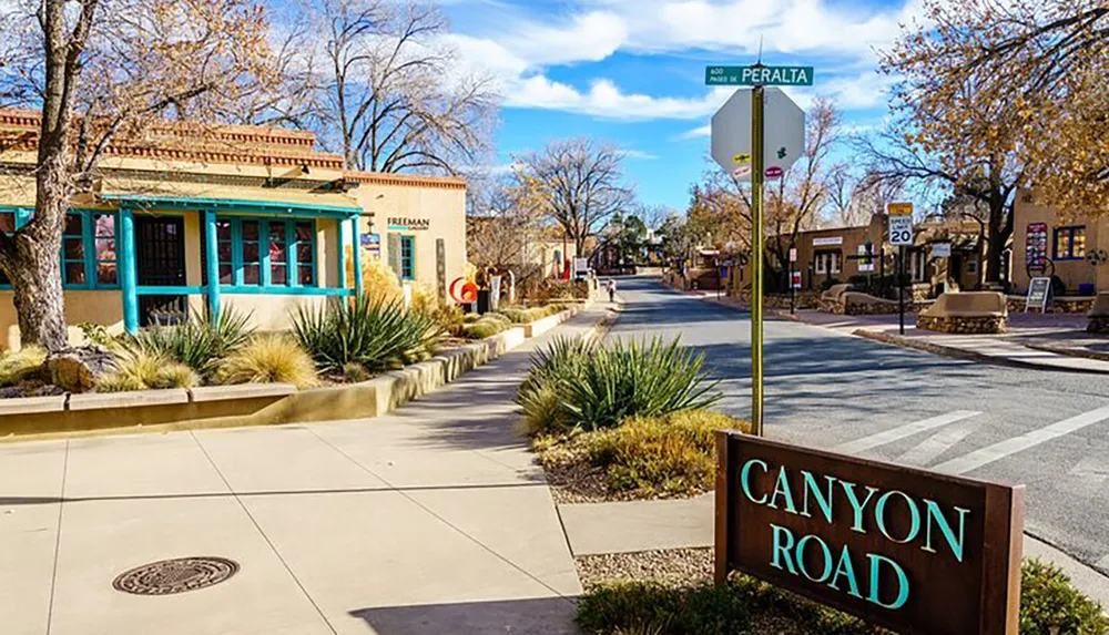 The image shows a sunny view of Canyon Road a street lined with Southwestern-style adobe buildings and galleries in a quiet and picturesque setting