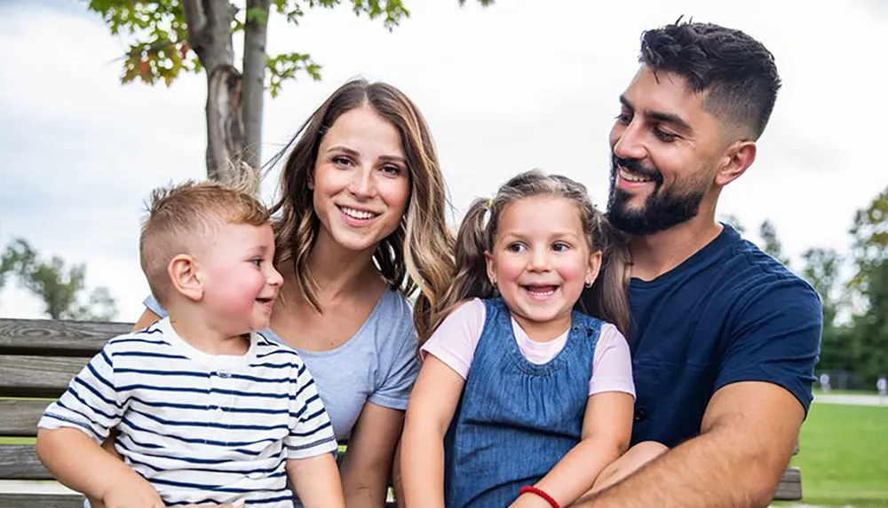 A happy family with two young children is sitting together outdoors smiling and enjoying a moment of bonding