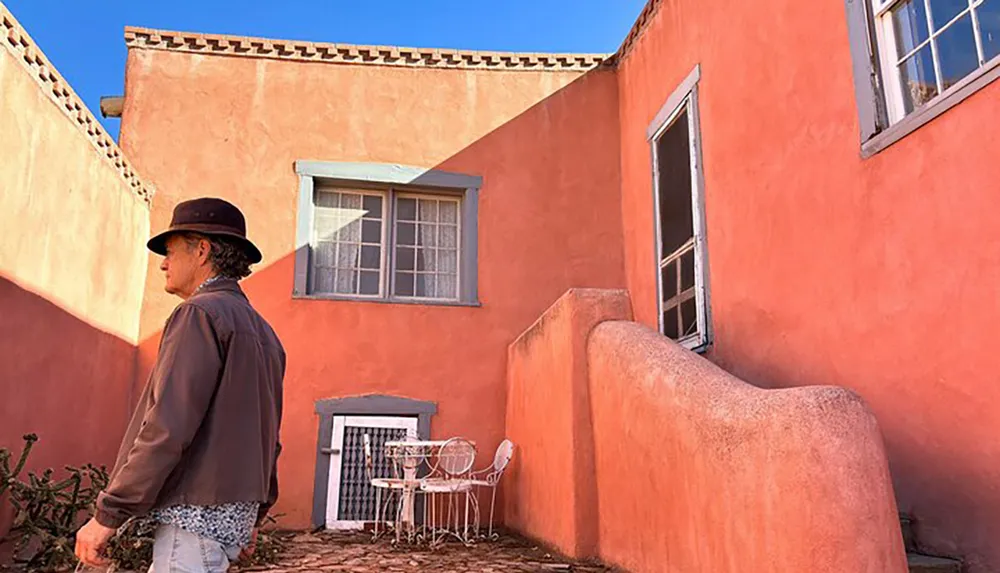 A person in a hat is walking past a salmon-colored building with distinctive Southwestern architecture under a clear blue sky