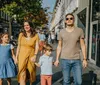 A family of four including a mother in a yellow dress a father in a brown t-shirt and two children is walking hand-in-hand along a sunlit urban sidewalk