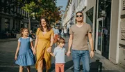 A family of four, including a mother in a yellow dress, a father in a brown t-shirt, and two children, is walking hand-in-hand along a sunlit urban sidewalk.