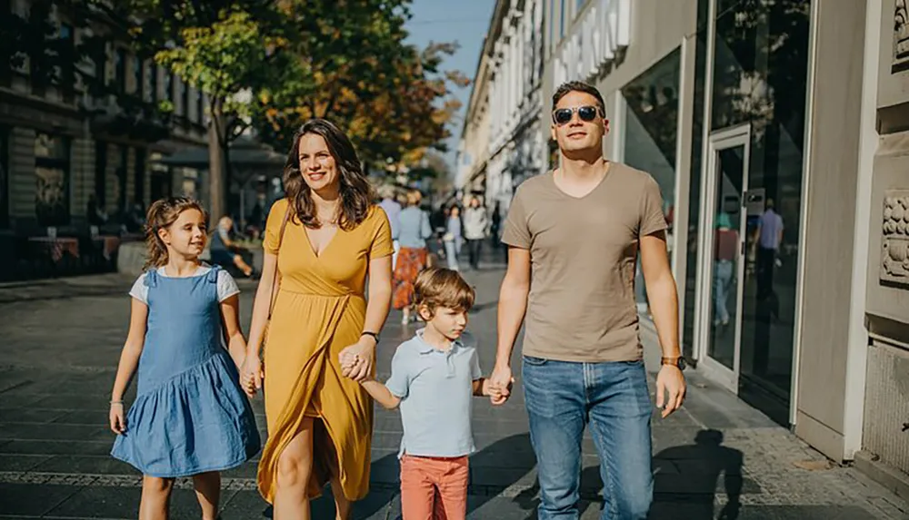 A family of four including a mother in a yellow dress a father in a brown t-shirt and two children is walking hand-in-hand along a sunlit urban sidewalk