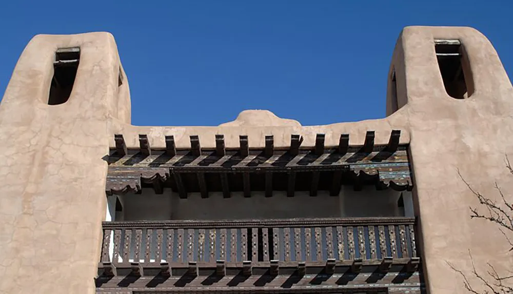 The image shows the upper section of a building with adobe-style architecture featuring a wooden balcony between two parapets under a clear blue sky