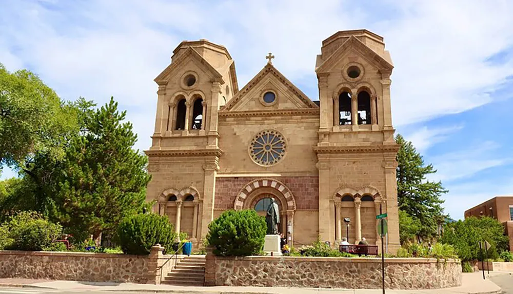 The image shows the symmetrical facade of a historic church with two bell towers a central rose window and an arched entrance set against a blue sky with greenery surrounding the structure