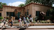 A group of people is walking past an adobe-style building adorned with sculptures, under a clear blue sky.