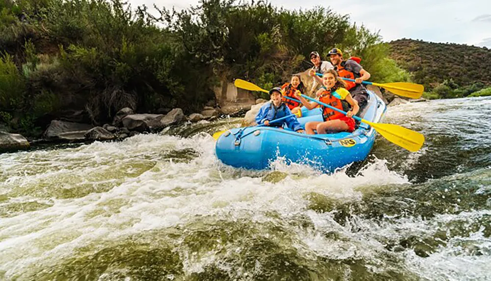 A group of people in life jackets are paddling a raft through white water rapids wearing expressions of excitement and concentration