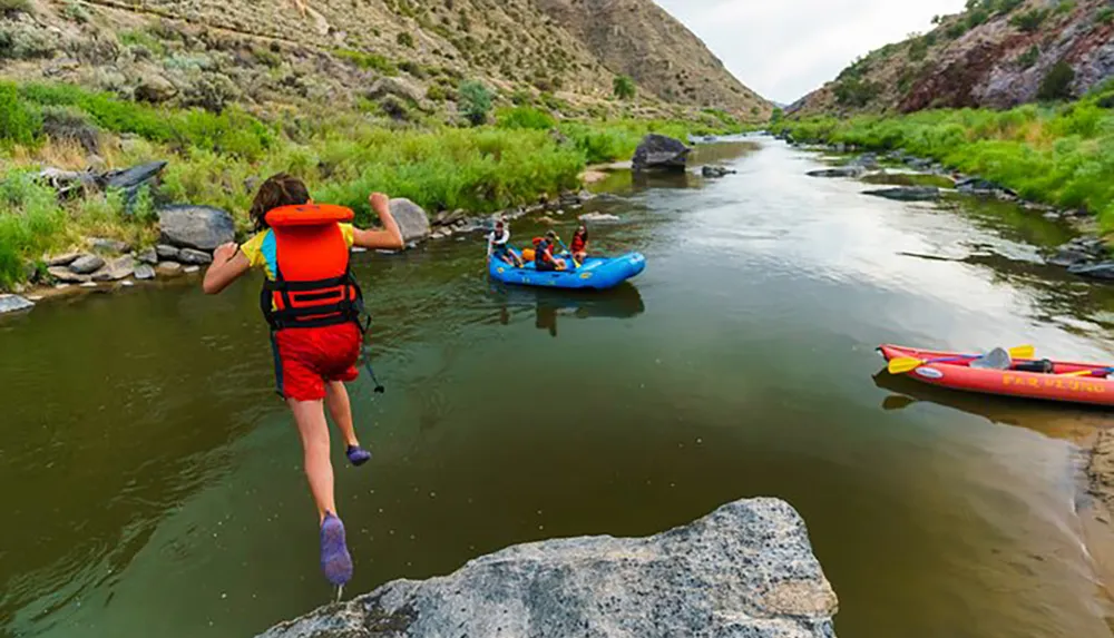 A person wearing a life jacket is jumping into a river where others are already aboard an inflatable raft and a kayak