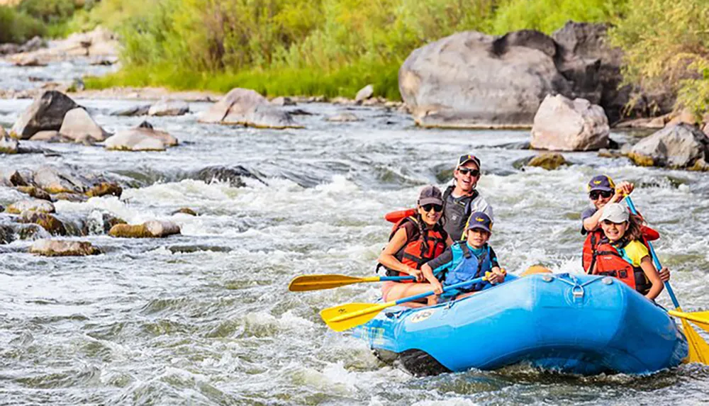 A group of people in life jackets and helmets are smiling as they navigate through rapids on a blue inflatable raft