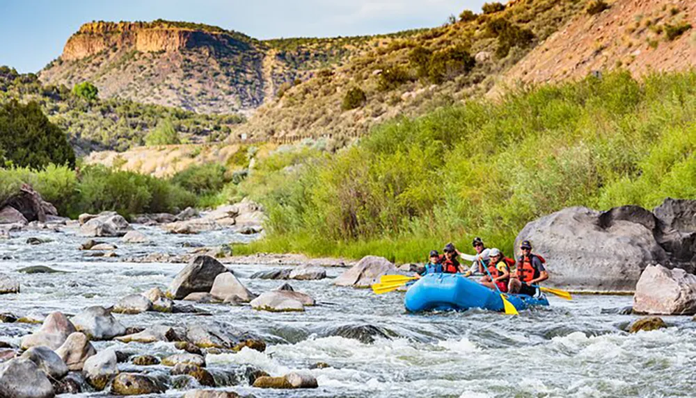 A group of people wearing life vests are rafting down a rocky river with lush greenery and a hill in the background