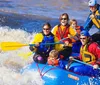 A group of excited people including children and adults wearing safety gear are white-water rafting in a turbulent river