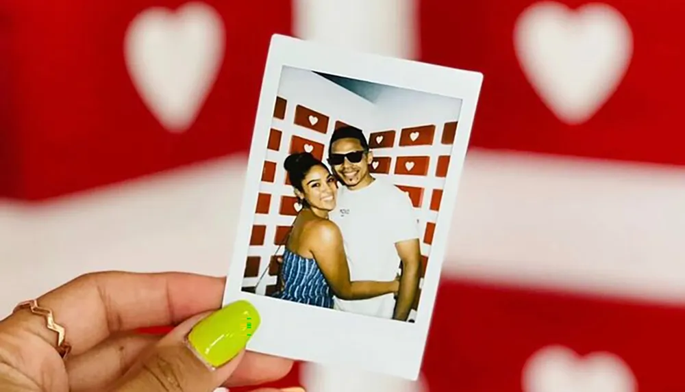 A persons hand is holding a Polaroid photo of a smiling couple posing together with a backdrop featuring heart designs