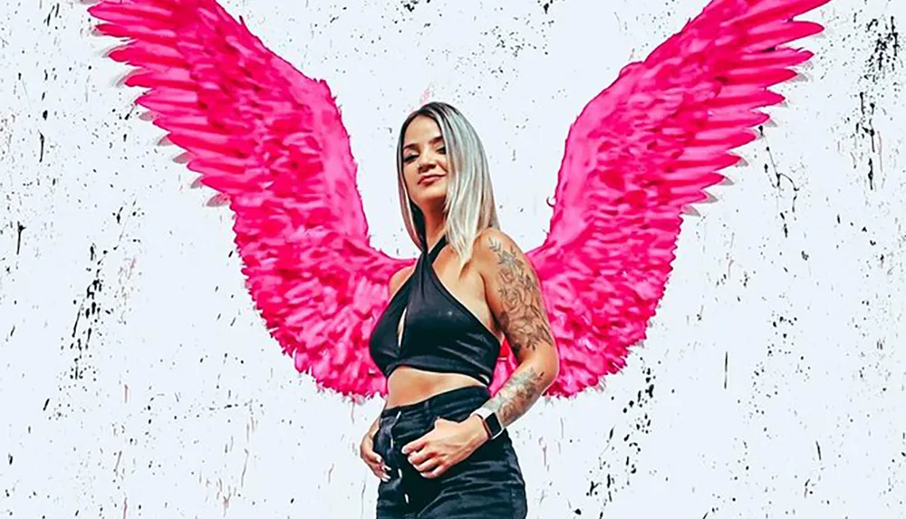 A person stands in front of a wall with a mural of vibrant pink wings creating the illusion that the wings are part of their own silhouette