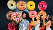 Three people are playfully interacting with large decorative doughnut designs on a wall.