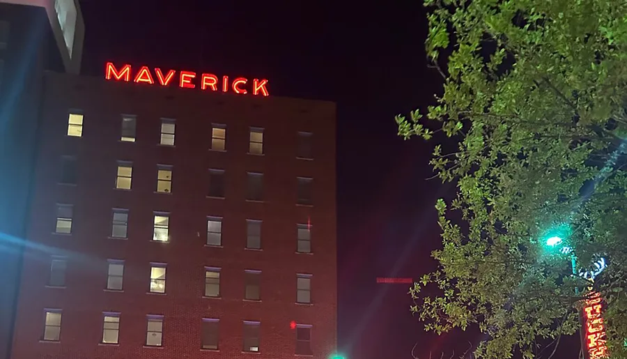 The image shows a nighttime view of a building with the red neon sign MAVERICK at the top, some illuminated windows, and the silhouette of a tree in the foreground.