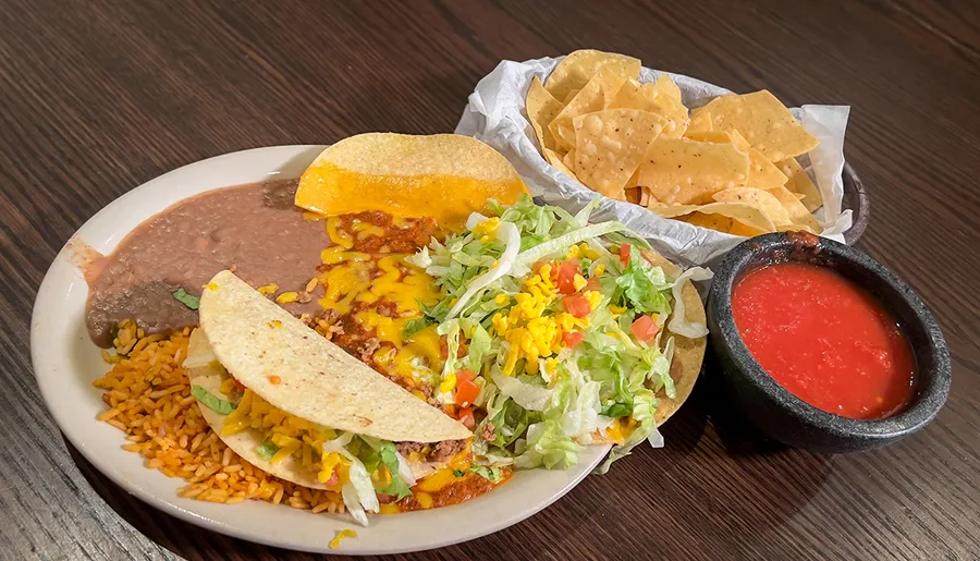 This image features a plate of Mexican food including tacos with side servings of rice, beans, tortilla chips, and salsa.