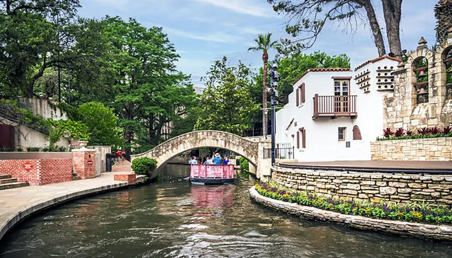 A picturesque scene of a riverwalk with people enjoying a boat tour under an arched bridge with charming white buildings and lush greenery surrounding the waterway.