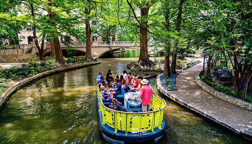 Tourists enjoy a guided boat tour along a lush tree-lined canal with pedestrian pathways and bridges in an urban setting