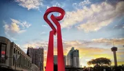 The image shows a large red abstract sculpture under a sky with a colorful sunset, with city buildings and a tower in the background.