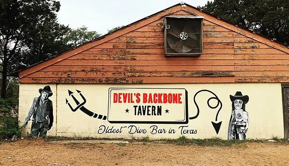 This is a photo of a rustic building with a mural declaring it the Devils Backbone Tavern claiming to be the Oldest Dive Bar in Texas adorned with stylized cowboy figures on its exterior walls