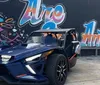 A blue three-wheeled Polaris Slingshot vehicle is parked on the side of a road next to a darker vehicle of the same model