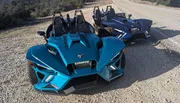 A blue three-wheeled Polaris Slingshot vehicle is parked on the side of a road next to a darker vehicle of the same model.