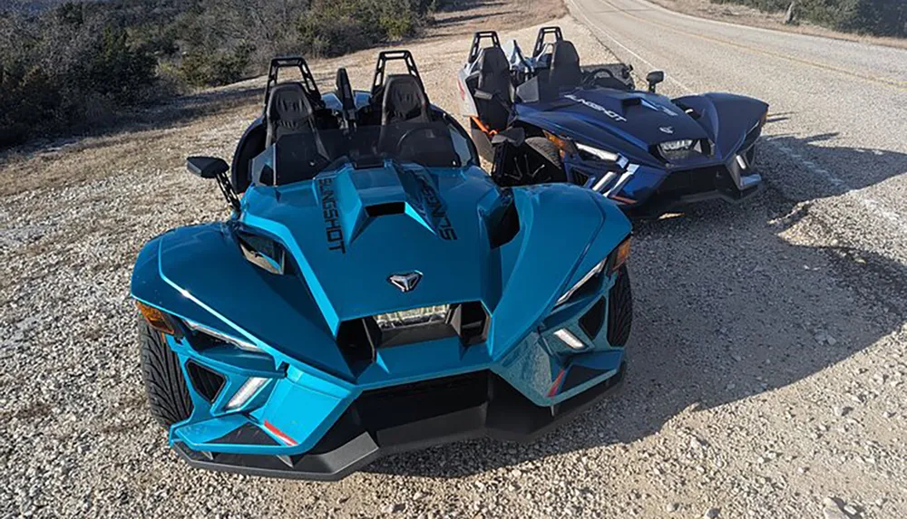 A blue three-wheeled Polaris Slingshot vehicle is parked on the side of a road next to a darker vehicle of the same model