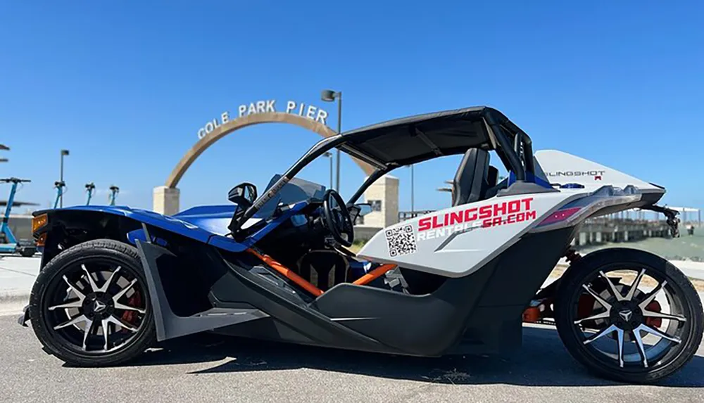 A blue and black Polaris Slingshot vehicle is parked by the Cole Park Pier with its distinctive three-wheeled design and open cockpit on display