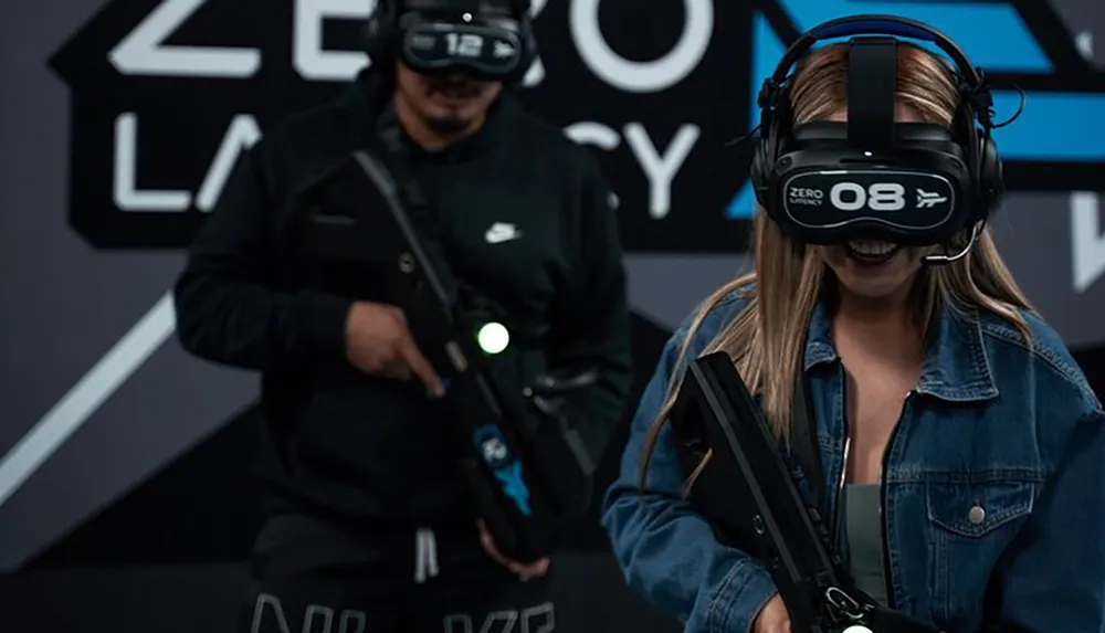 Two people are wearing virtual reality headsets and holding mock rifles presumably engaged in a VR gaming experience