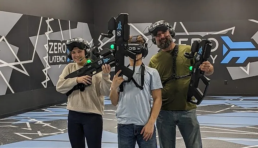 Three people are posing and smiling in a virtual reality gaming arena holding VR guns and wearing VR headsets