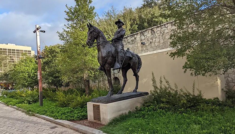 The image shows a bronze statue of a person mounted on a horse placed in a park setting with greenery and urban structures in the background