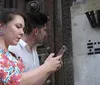 A woman and a man are standing close to a wall with the woman focused on her mobile phone and an enigmatic black symbol on the wall behind them