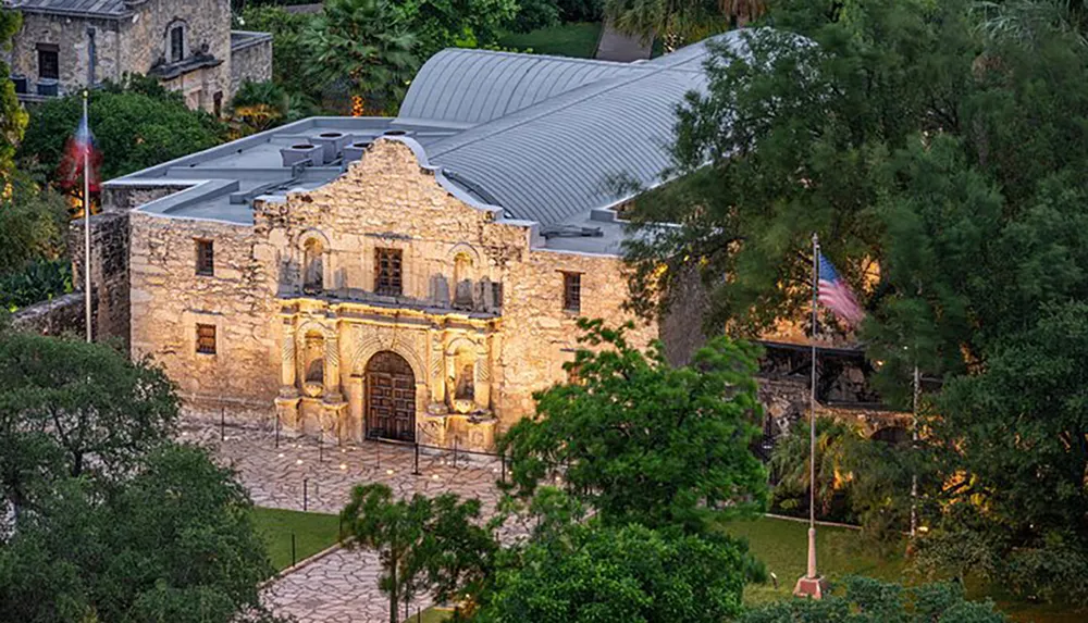 The image shows the historic Alamo mission at twilight with lights illuminating the facade and an American flag flying at its side