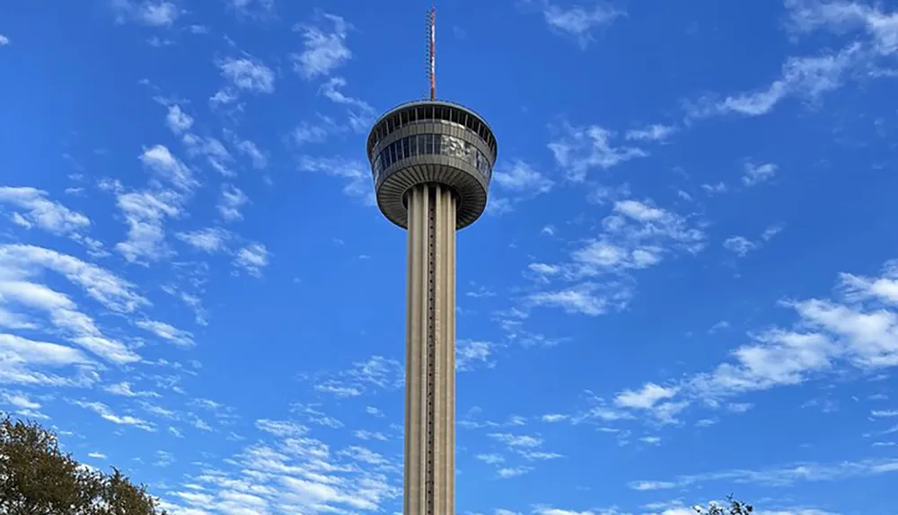 The image shows a tall observation tower with a circular viewing platform against a backdrop of blue sky dotted with white clouds