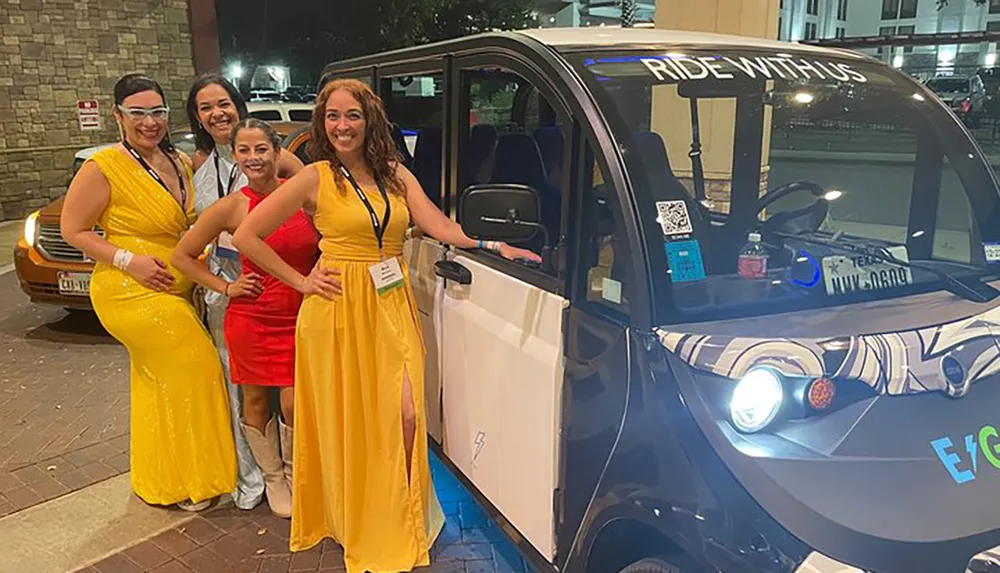 Four women in formal attire are posing with smiles beside a small modern electric vehicle at an evening event