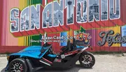 A blue and black three-wheeled vehicle is parked in front of a colorful mural that spells out 