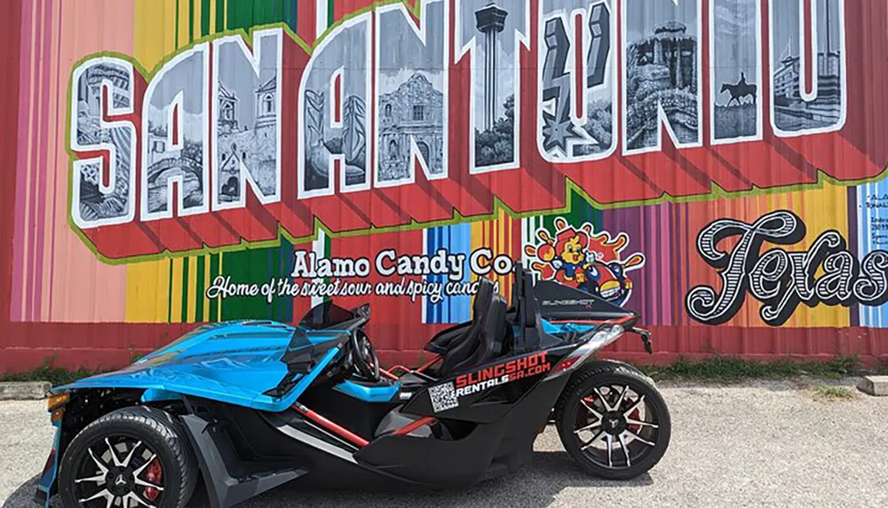 A blue and black three-wheeled vehicle is parked in front of a colorful mural that spells out SAN ANTONIO TEXAS