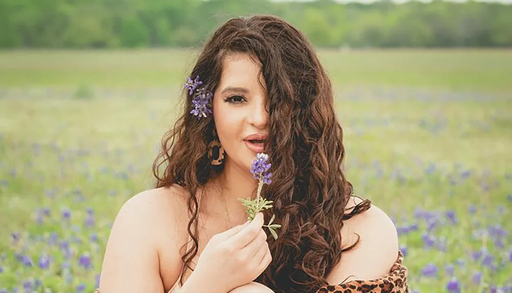 A woman with flowers in her hair is holding a bluebonnet posing in a field with more bluebonnets in the background