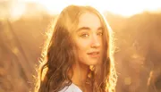 A young woman with wavy hair is backlit by a warm, golden sunset, creating a soft, glowing portrait amidst a blurred natural background.